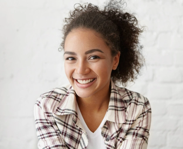 Young woman smiling at camera wearing plaid shirt in bright clinical setting.