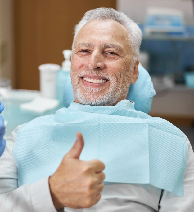 Smiling senior man giving thumbs up in dental chair wearing protective bib at dentistry office.