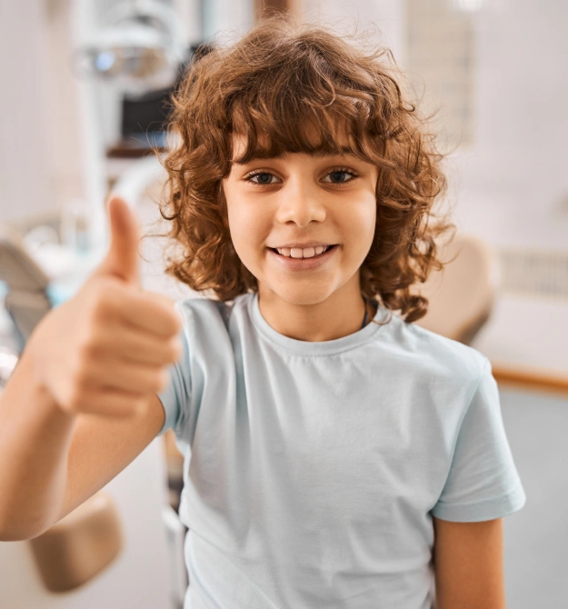 Happy child with curly hair giving thumbs up in dental office setting.