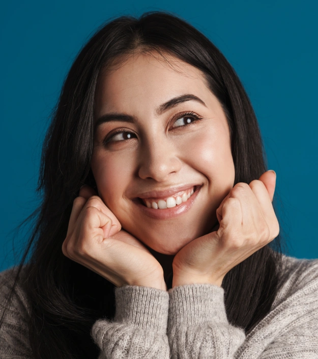 Young woman smiling at camera, hands on face, blue background, dental office portrait.