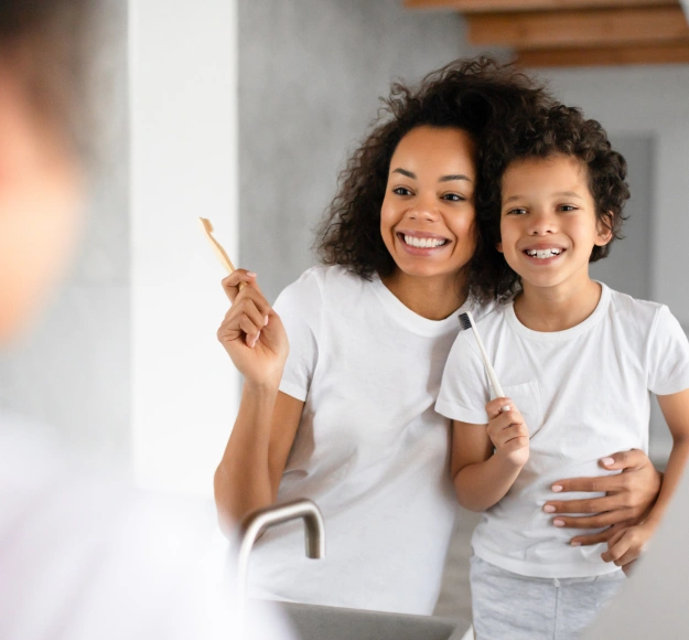 Mother and child brushing teeth together at bathroom sink, smiling.