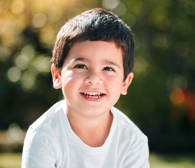Happy young boy smiling outdoors in white shirt, sunny garden background.
