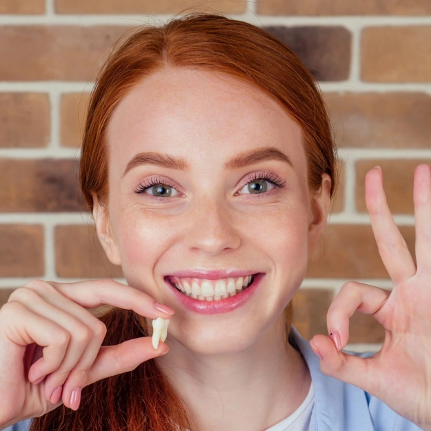 Young woman with red hair smiling, pointing to teeth against brick wall background.
