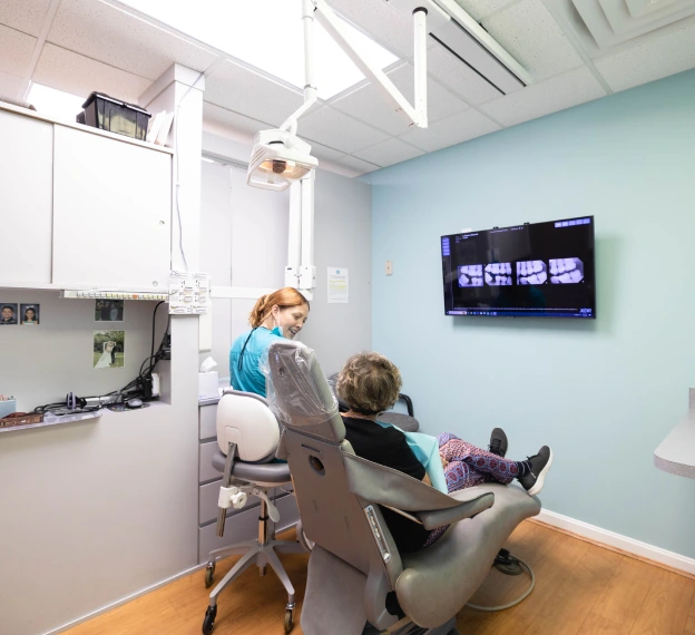 Dental hygienist treating young patient in modern clinic operatory with mounted monitor.