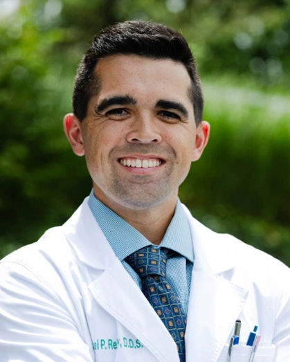 Professional male dentist in white coat and blue tie smiling outdoors with green background.