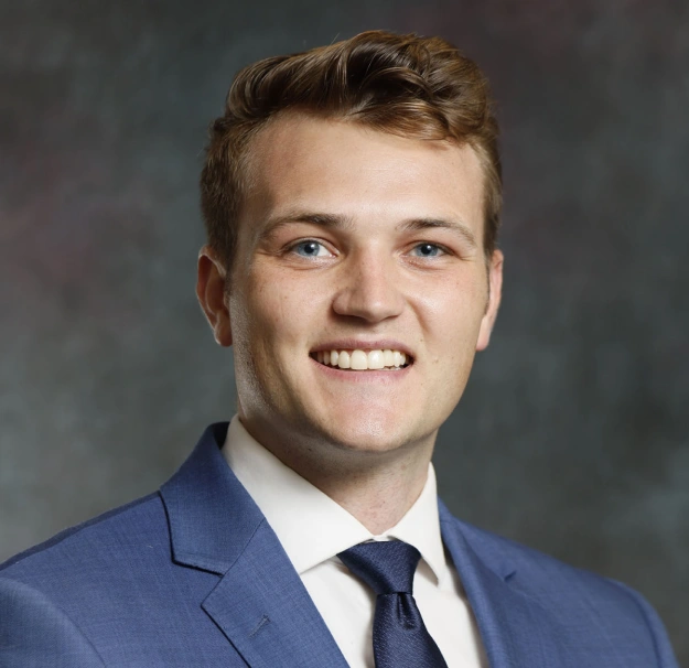 Professional headshot of smiling man in blue suit and tie against gray background.
