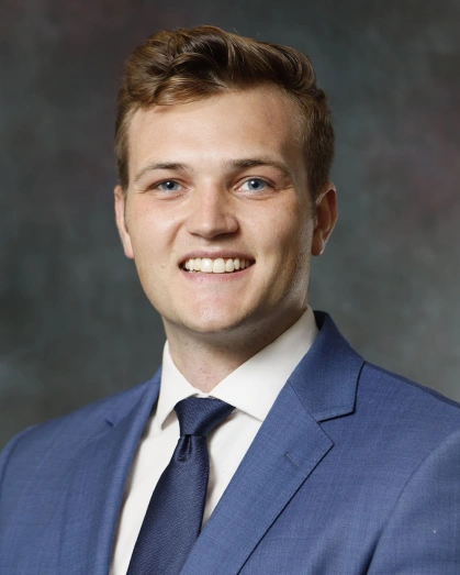 Professional headshot of smiling man in blue suit and tie against gray background.